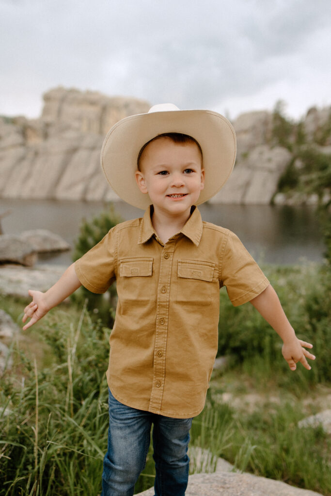 Little boy in cowboy hat with arms out while dancing. Sylvan Lake in Custer State Park, SD is behind him.